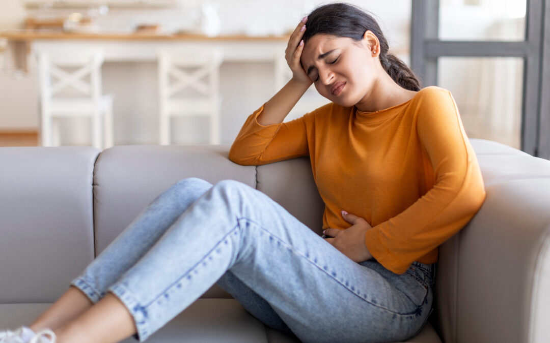 woman in orange shirt and jeans sitting on couch holding her stomach and head in pain, showing symptoms of food poisoning or a stomach virus.