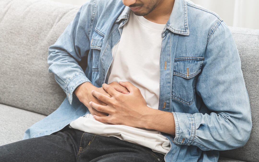 Man with jean jacket, white shirt, and jeans seated on a couch holding his abdomen in visible discomfort, showing signs of stomach or upper abdominal pain possibly related a norovirus infection.